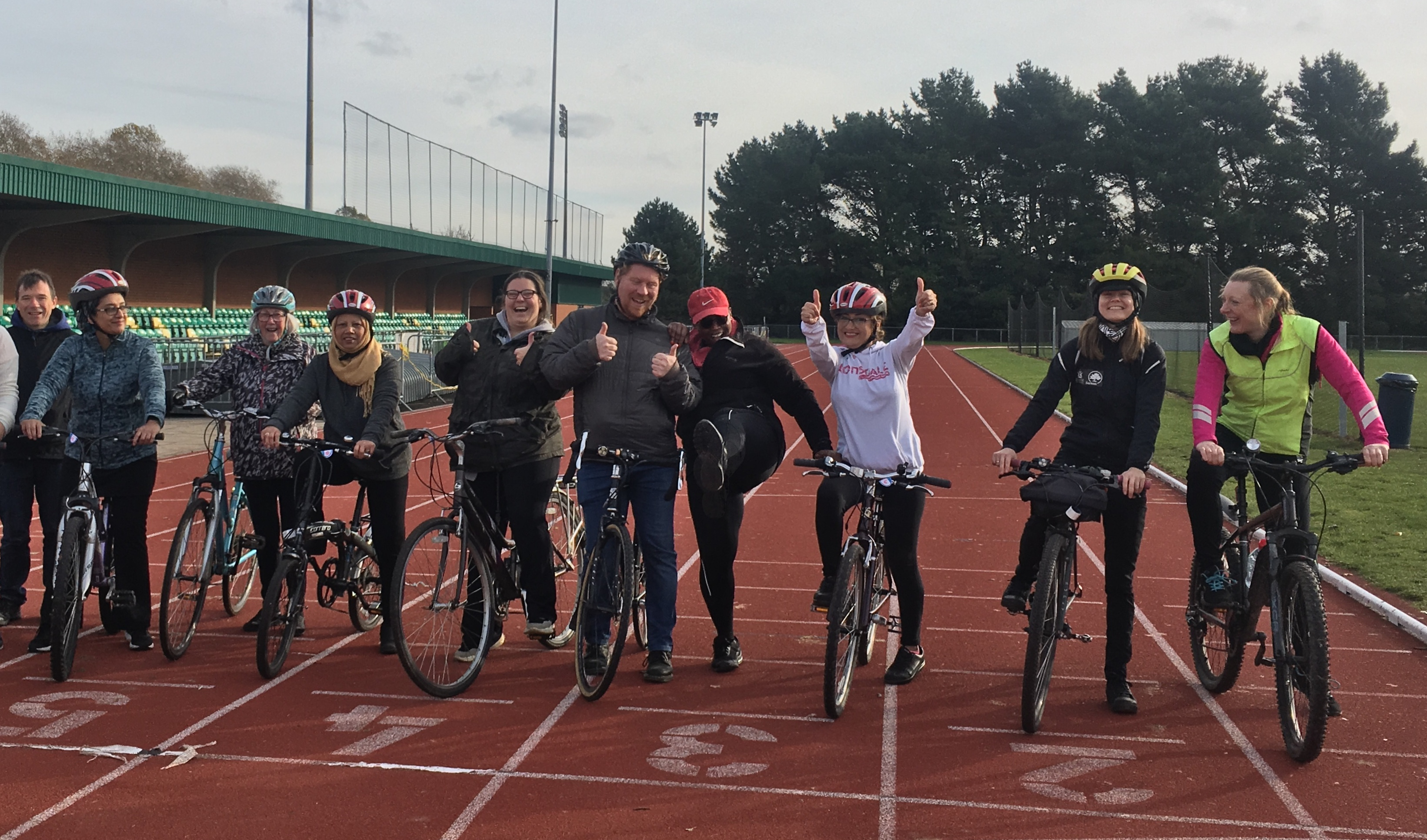 A group of 10 adult cyclists in helmets pose with their bikes on an athletic track
