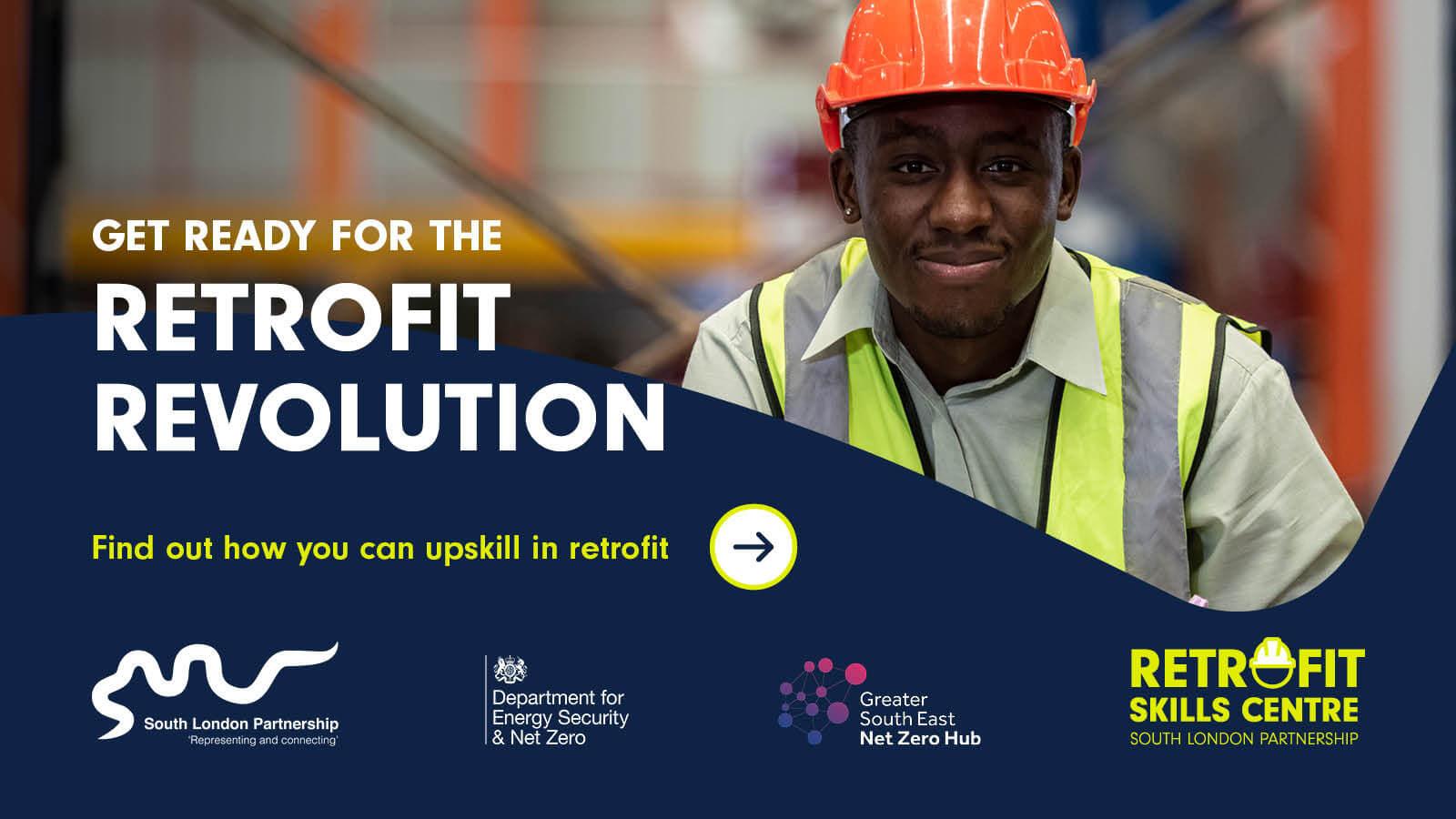 A man in an orange hard hat and hi-vis vest smiles for the camera. In the background is a construction site.