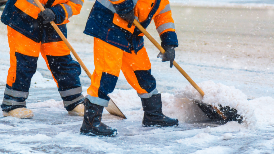 Two workers in gloves, boots and orange overalls clear snow and ice from a pavement with shovels