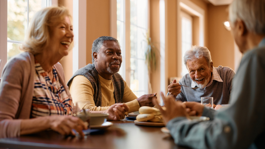 Three men and a woman, all in their 60s or 70s, sit at a table eating and laughing