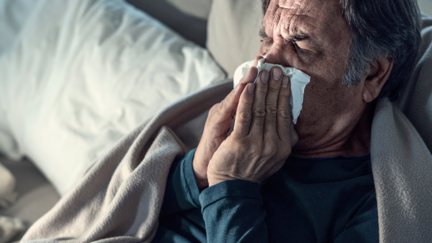 An older man with grey hair blows his nose while sitting up in bed