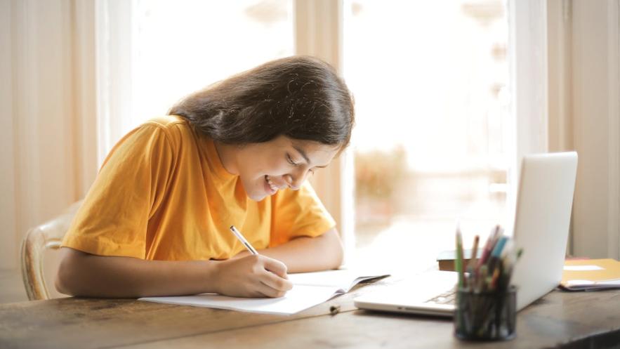 A smiling young woman sits at her desk in a sunny room, making notes. A laptop is open before her.
