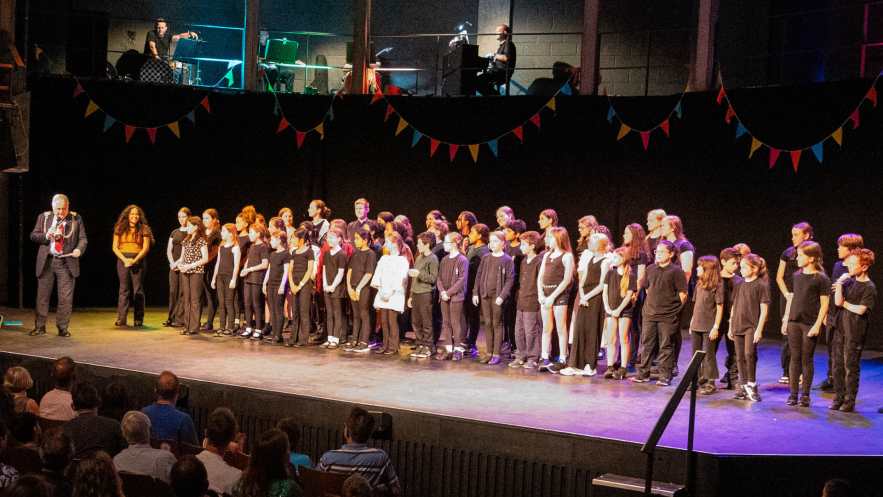 On A theatre stage the mayor of Kingston stands to the left of the young ensemble cast and choir of Bridging Kingston: A Musical Odyssey   