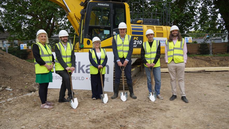 Image of six people wearing high visibility jackets and hard hats, holding spades in front of construction machinery. 